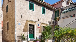 A stone building with a green door, a small garden with plants, and a paved street. - Sun Spalato Mak Apartments (Split)