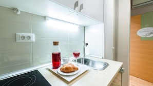 Ett modernt kök med en diskho i rostfritt stål, en vit bänkskiva och en svart induktionshäll. På bänkskivan står en flaska röd dryck, ett glas rött vin och en croissant.