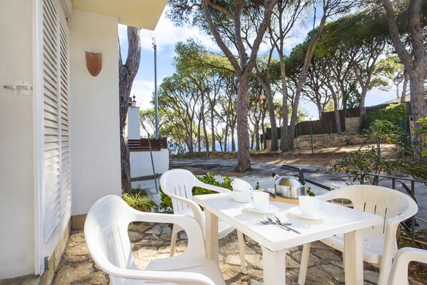 Een terras met witte plastic stoelen en een eettafel, met uitzicht op een natuurlandschap met bomen en zee.