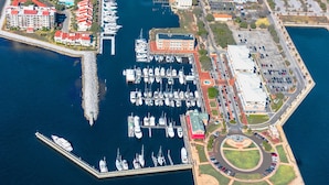 An aerial view of a marina with multiple boats docked, a large building with a red roof, and a parking area with cars.