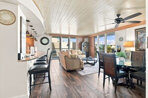 A spacious living area with a high ceiling, a dining area with a glass table, and a view through large windows.