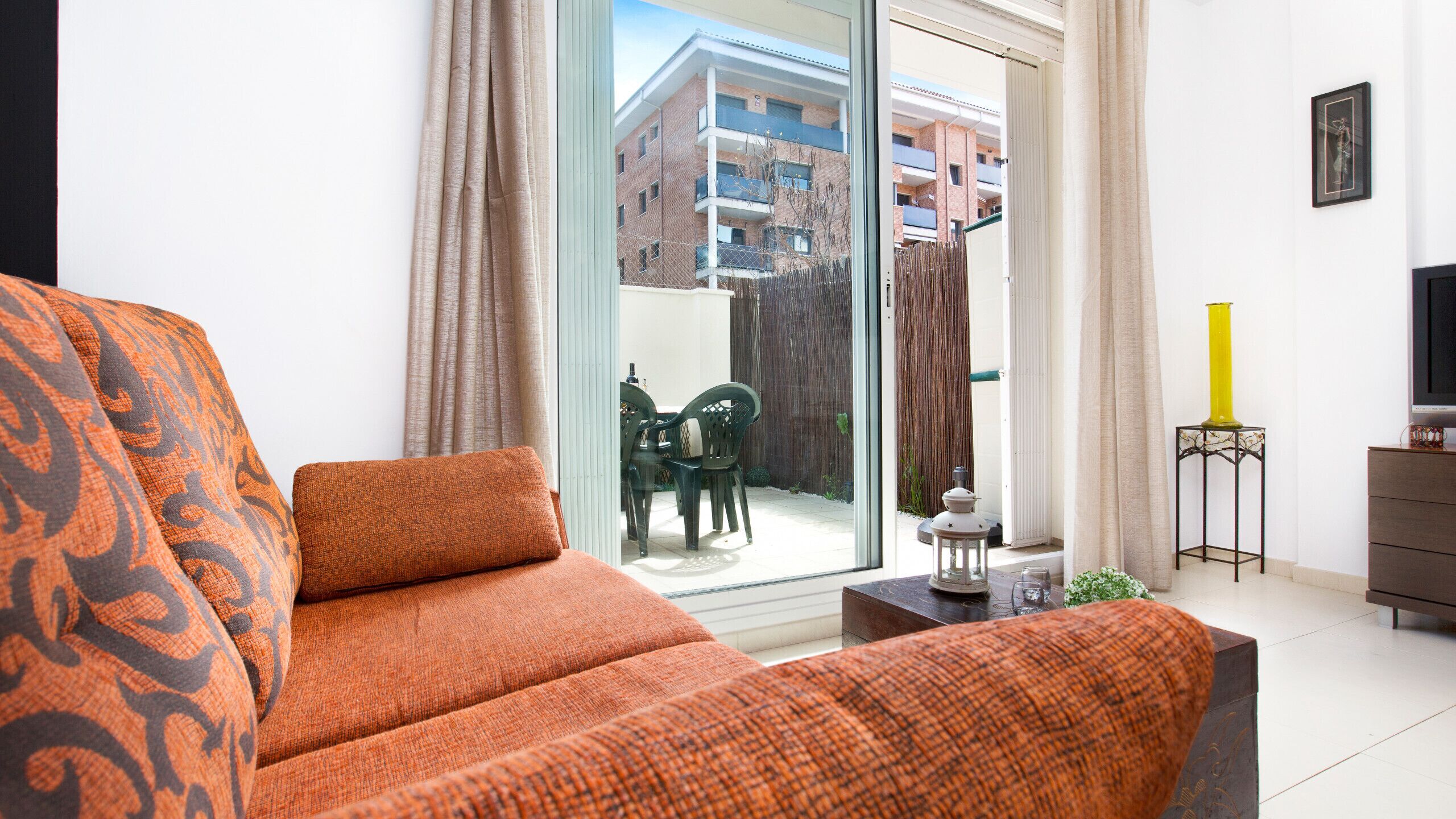 A modern living room with a sofa, a TV, and a view of a balcony with outdoor furniture.