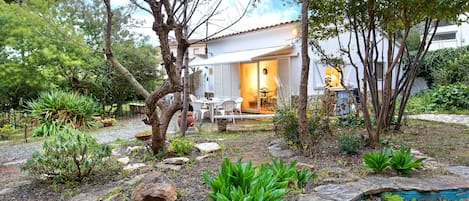 Una casa con un patio, rodeada por un jardín con diversas plantas y un pequeño estanque.