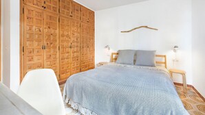Une chambre avec une grande armoire en bois, un lit recouvert d’une couverture à motifs, deux lampes de chevet et un simple luminaire fixé au mur.