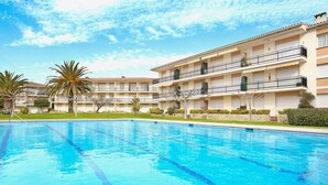 A swimming pool with clear blue water, surrounded by a multi-story building with balconies and a garden area with palm trees. - 1CB K2 -2 Bedroom apartment in a very quiet area with garden and communal pool near the beach of Calella de Palafrugell (Calella de Palafrugell)