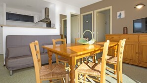 Uma sala de jantar com mesa e cadeiras de madeira, um sofá cinzento e uma cozinha com armários e fogão.