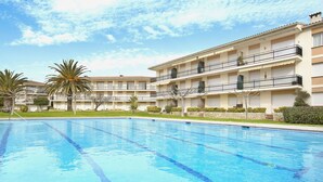 Piscina com água azul cristalina, rodeada por um edifício com vários andares e varandas, e um jardim com palmeiras.