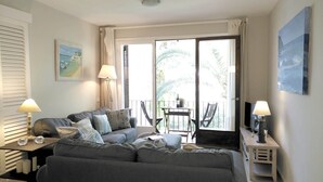 A modern living room with a grey sofa, a small table, and a TV. There is a balcony with a table and chairs, and a painting on the wall.