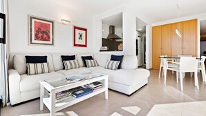 A modern living room with a white sofa, striped pillows, a glass coffee table with books, and a dining area with white chairs and a wooden cabinet.