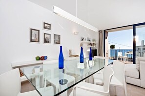 A modern dining area with a glass table and white chairs, a balcony with a view, and a white interior with framed pictures on the wall.