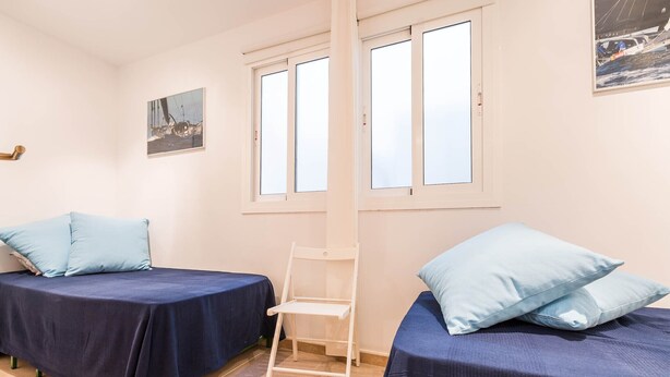 A room with two beds, each with a blue bedspread and matching pillows, a white chair, and a window with white frames.