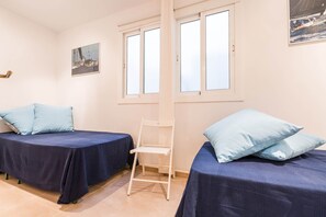 A room with two beds, each with a blue bedspread and matching pillows, a white chair, and a window with white frames. - 1SR 01 - Beautiful and spacious new build apartment with sea view located in  the second row of houses lining the sea in Llafranc. (Llafranc)