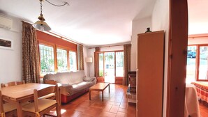 A living room with a sofa, dining table, chairs, and a lamp. There are windows with curtains and a door leading outside. - 1AL 01 - Simple apartment with terrace near the beach of Llafranc (Llafranc)