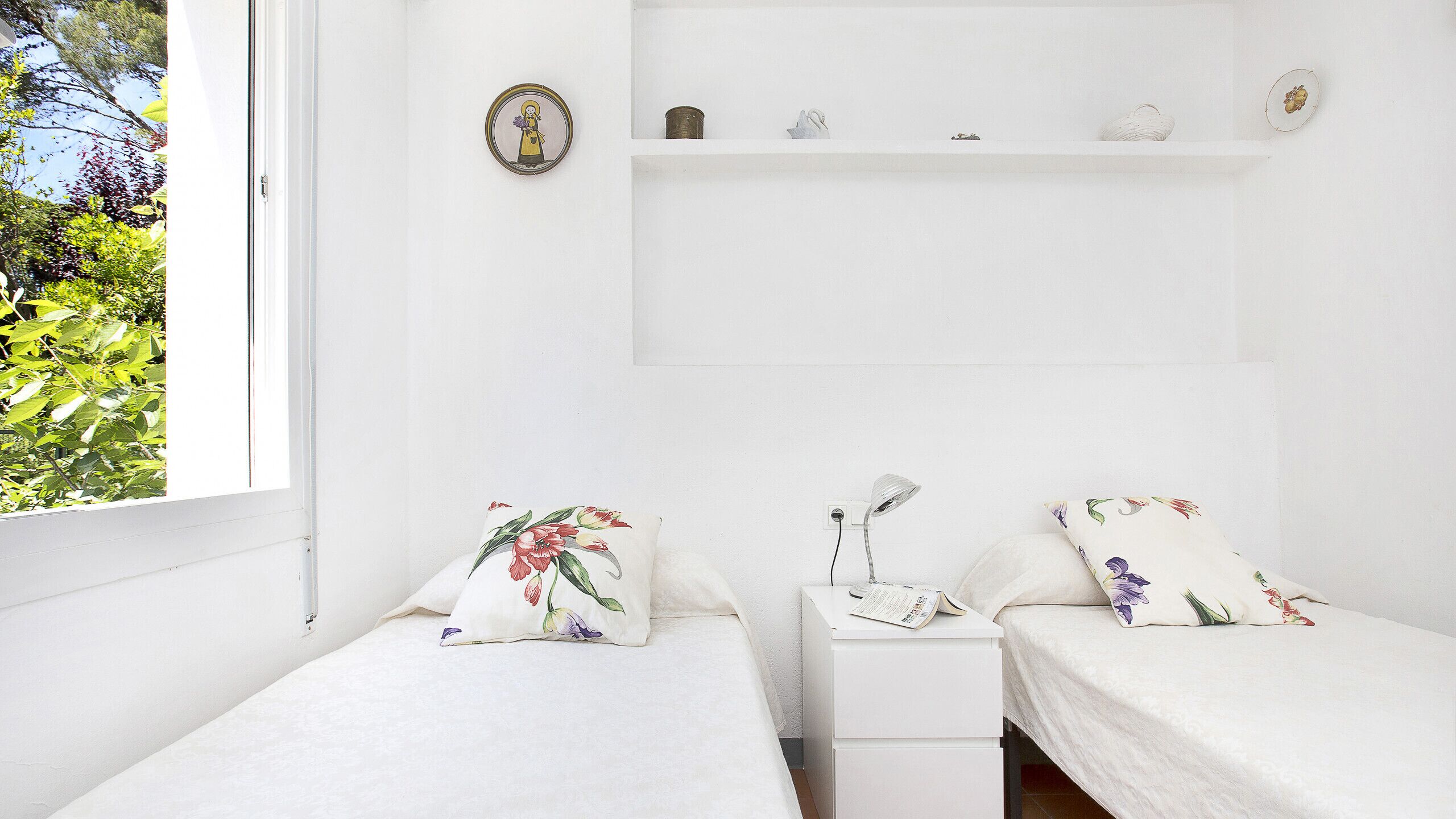 Une chambre à coucher au style minimaliste, avec deux lits, une table de chevet, une étagère ornée d’objets décoratifs et une fenêtre donnant sur un paysage verdoyant.