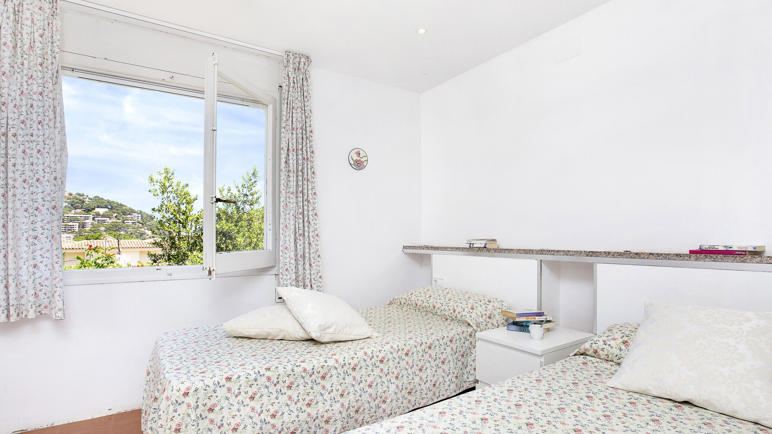 Une chambre avec deux lits, une fenêtre avec des rideaux, une table de chevet avec des livres et une vue sur un paysage verdoyant.