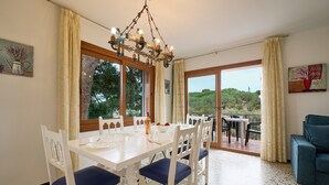 A dining area with a white table and blue chairs, a chandelier, and a view through large windows. - 1BENET 1 - House divided into 3 totally independent apartments with shared pool just 1 km from the beach of Calella de Palafrugell (Calella de Palafrugell)