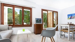 A modern living room with a sofa, a dining table, chairs, a TV on a wooden stand, and large windows with a view of trees and a building.