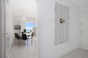 Un espace de repas moderne avec une table et des chaises blanches, un luminaire suspendu et une vue sur la mer grâce à de grandes fenêtres.