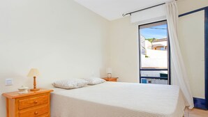 Un dormitorio con una cama, una mesita de noche de madera, una lámpara y una ventana con vistas a edificios.