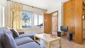 Un saló modern amb un sofà, una taula amb llibres, una televisió i una porta de fusta que dona a un balcó amb vista al mar.