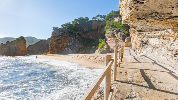 A coastal walkway with a wooden railing, leading to a sandy beach and rocky cliffs. - 1OP 01 - Apartment located in Pals, in a residential complex with swimming pool, 400 m from the beach (Pals)