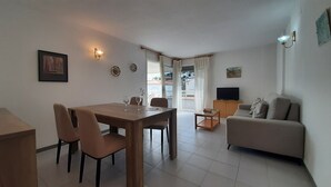 A living room with a dining table, chairs, a sofa, a TV, and a balcony with curtains. - 1OP 01 - Apartment located in Pals, in a residential complex with swimming pool, 400 m from the beach (Pals)