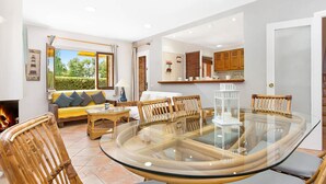 Uma sala de estar espaçosa com uma mesa de jantar de vidro rodeada por cadeiras de vime, um sofá com almofadas e vista para um jardim através de grandes janelas.