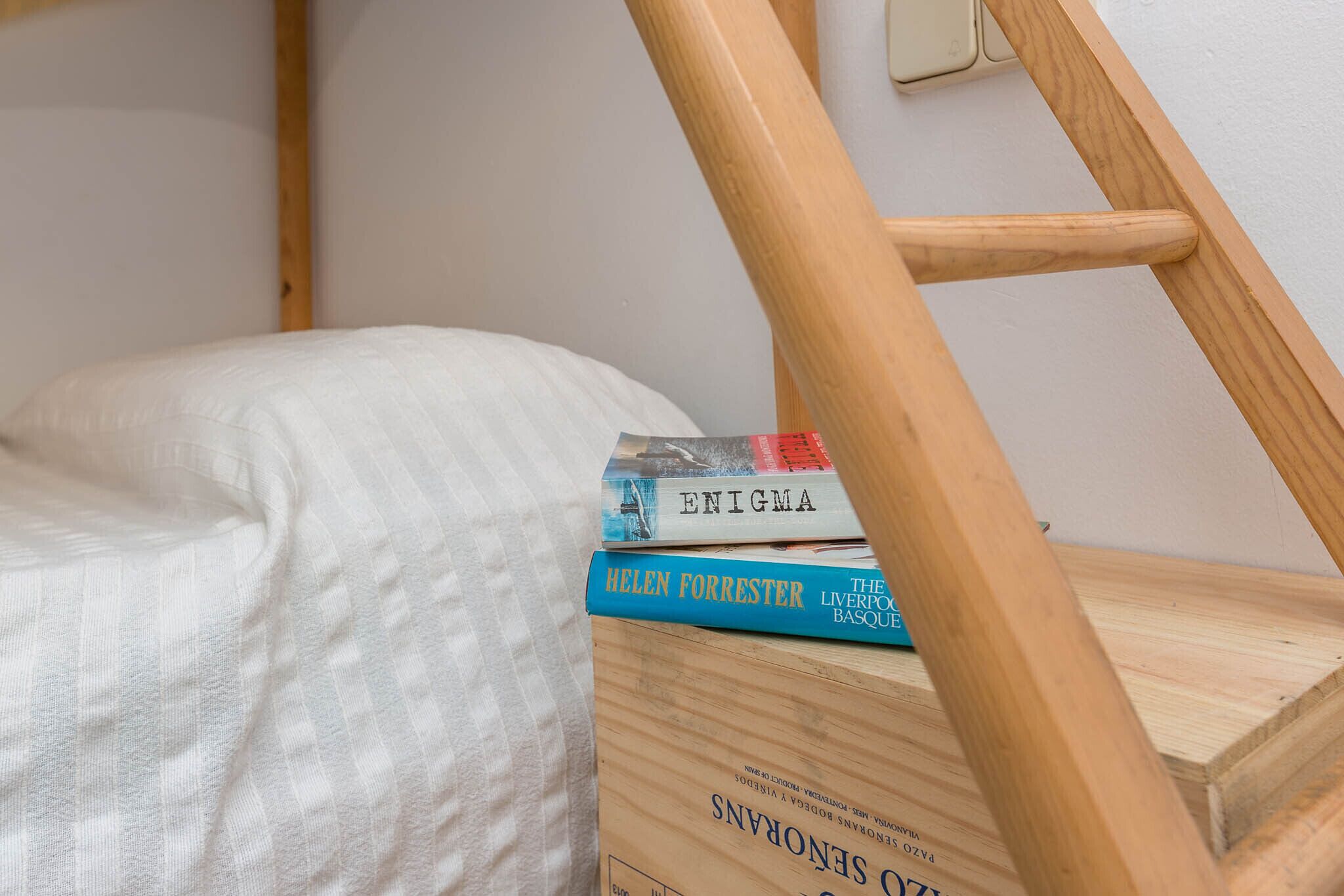 A bunk bed with a wooden ladder, a white mattress, and a stack of books on the bed.