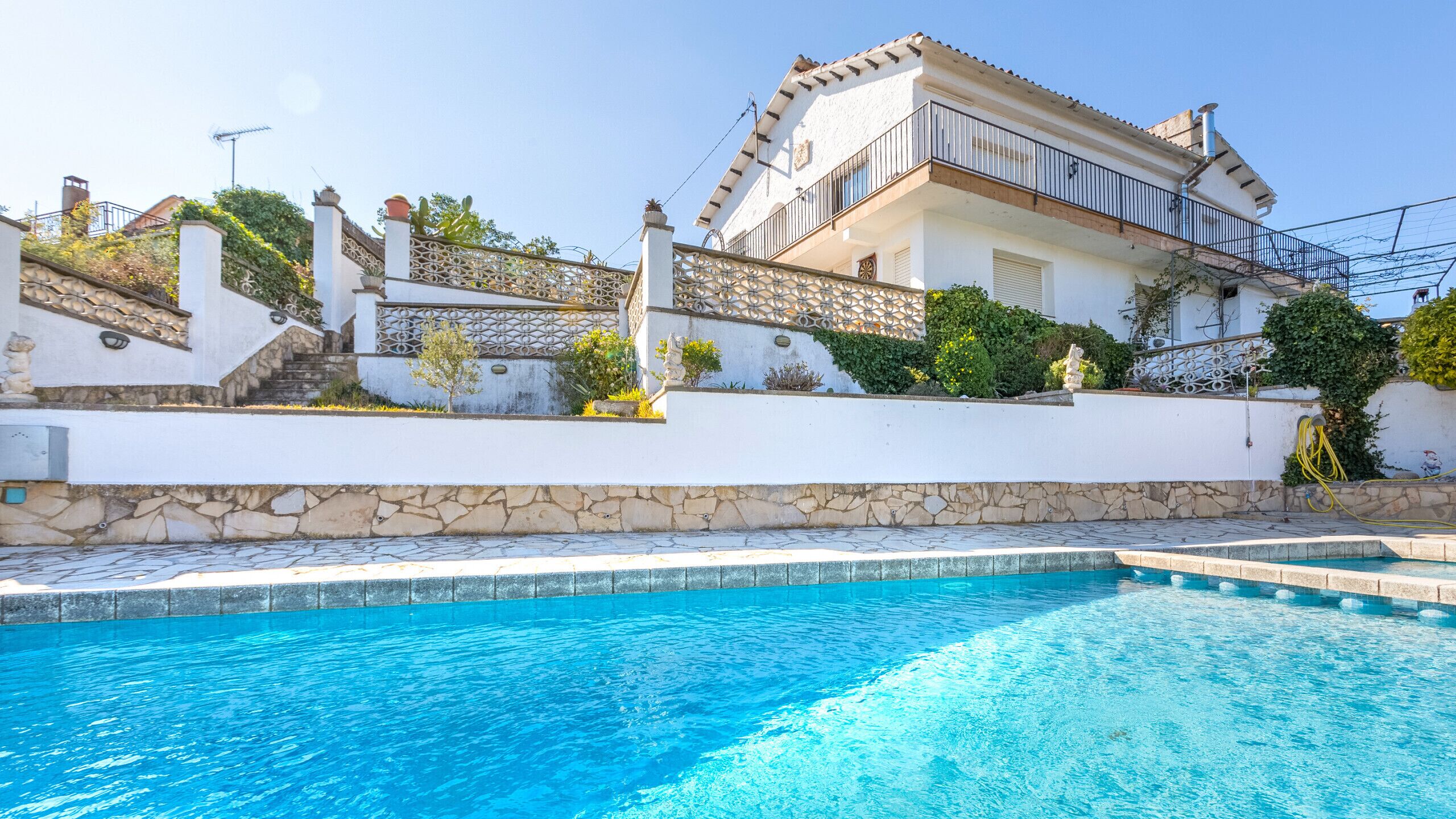 Blick auf einen Pool vor einer weißen Villa mit Mauerwerk, einer Treppe und einem Balkon.