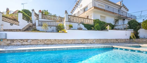 A poolside view of a white villa with a stone wall, a staircase, and a balcony.