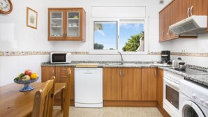 Cocina compacta con armarios de madera, microondas, lavavajillas, lavadora y ventana con vistas al verdor.