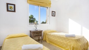 Una habitación con dos camas, una mesita de noche de madera, una lámpara y un ventanal con vistas a árboles y un edificio.