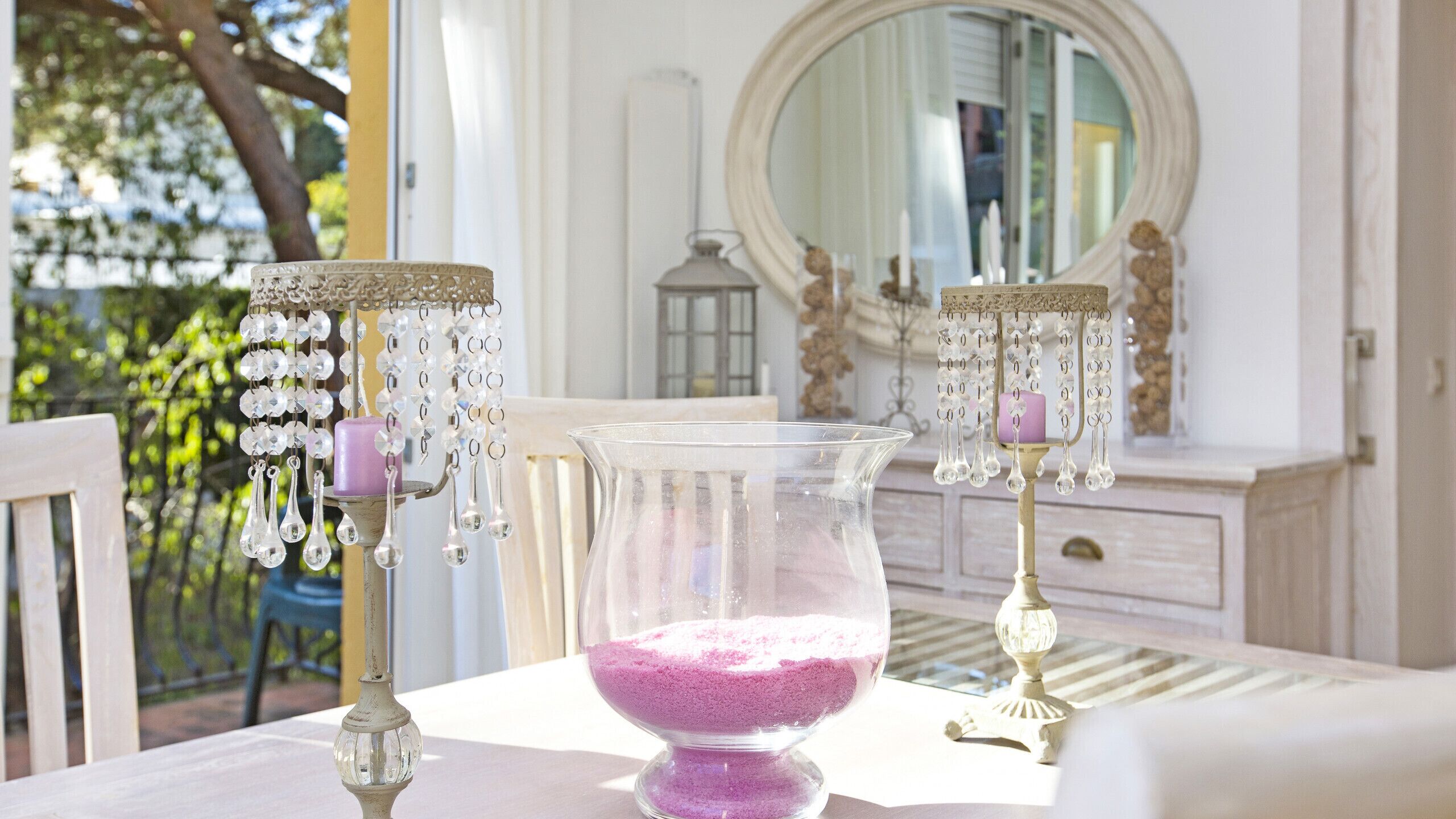 Una mesa con un florero de vidrio transparente que contiene arena rosa, dos candelabros ornamentados con detalles de cristal, y un espejo que refleja una habitación con un tocador y una ventana.