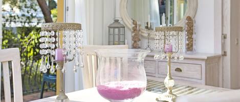Una mesa con un florero de vidrio transparente que contiene arena rosa, dos candelabros ornamentados con detalles de cristal, y un espejo que refleja una habitación con un tocador y una ventana.