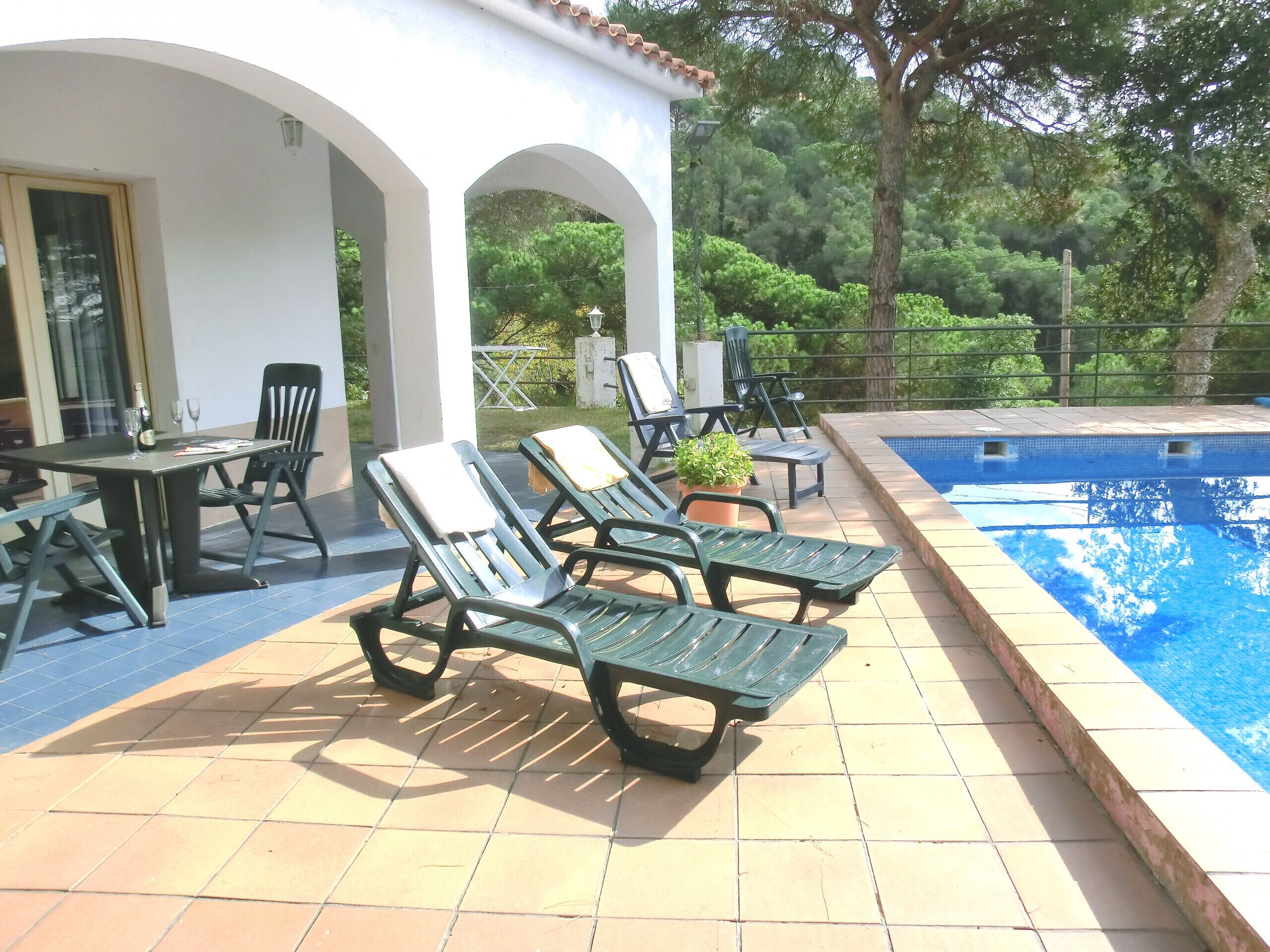 Zona de piscina con sillones de descanso, una mesa y una puerta de cristal que da a un porche cubierto.