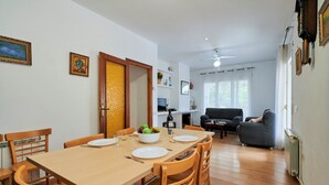 A dining area with a wooden table set for four, surrounded by wooden chairs. A bowl of fruit and a bottle of wine are on the table. There is a sofa and a television in the background. - 2SEN01 - House with private pool located in a quiet area just 2.5 km from the beautiful beach of Canyelles (Lloret de Mar)