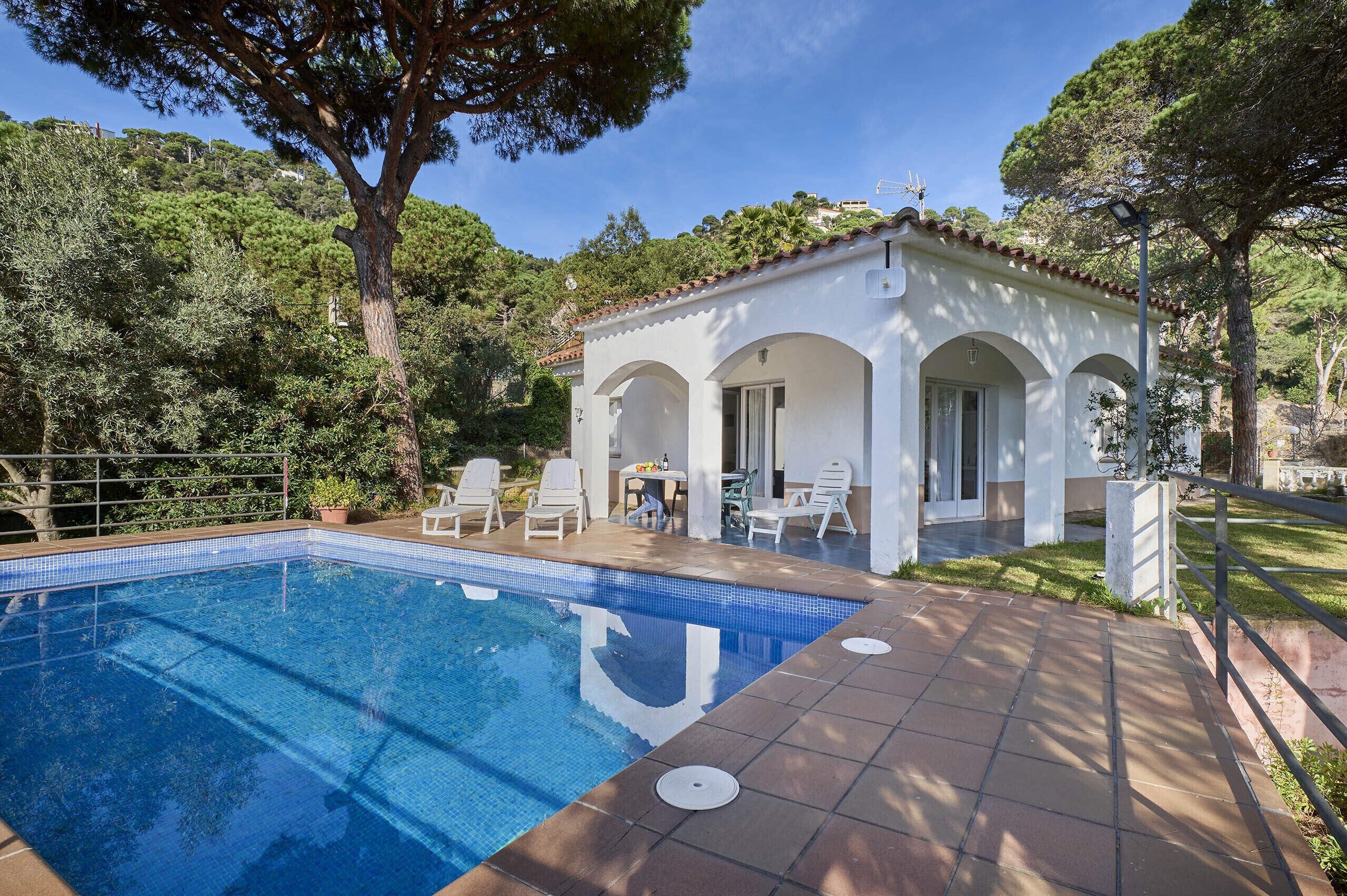 A poolside area with a clear blue pool, white lounge chairs, and a small building with arches.