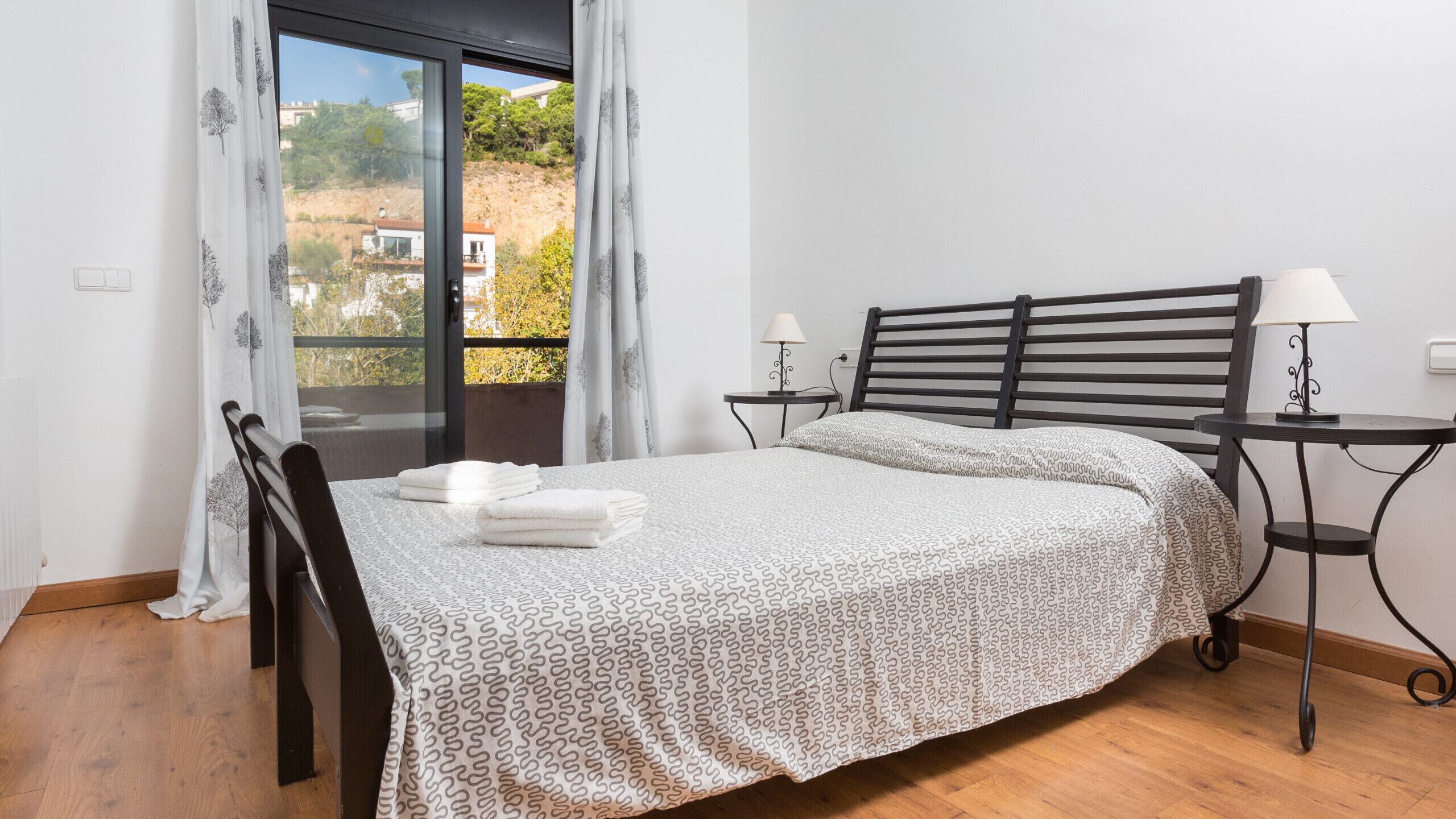 A bedroom with a bed, two bedside tables, a chair, a window with curtains, and a view of buildings and trees.