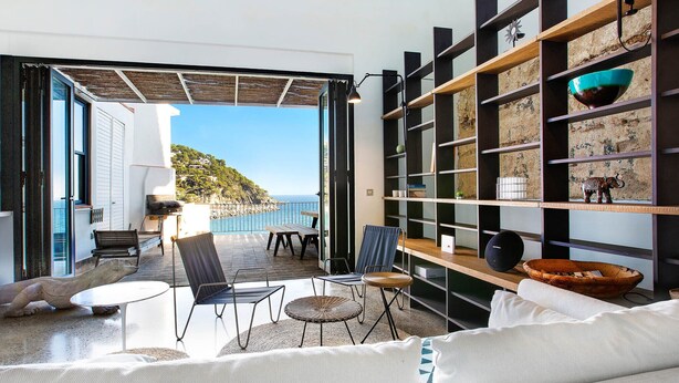 A modern living room with a large white sofa, a coffee table, and a dining area with chairs and a table. There is a bookshelf with decorative items and a view of the sea through an open door. - 1MIRAD 01 - Beautiful house renovated with great taste, with fantastic views of the sea and direct access to the beach of Llafranc (Llafranc)
