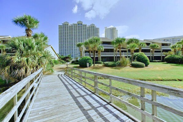 A wooden boardwalk leading to a building complex with multiple floors and a grassy area with palm trees.