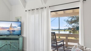 A living room with a television displaying a sailboat on the water, a wooden rocking chair, and a view of a lake through a balcony.