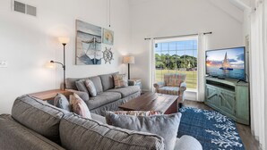A living room with a large sofa, a coffee table, and a television. There is a window with a view of the outdoors.