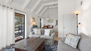 A spacious living area with a wooden coffee table, grey sofas with striped cushions, a patterned rug, and a view through a glass door to an outdoor deck.