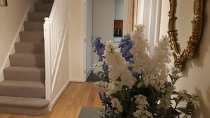 Uma escadaria com degraus revestidos de carpete bege, corrimão branco e piso de madeira. Há um vaso com flores brancas e azuis à direita.