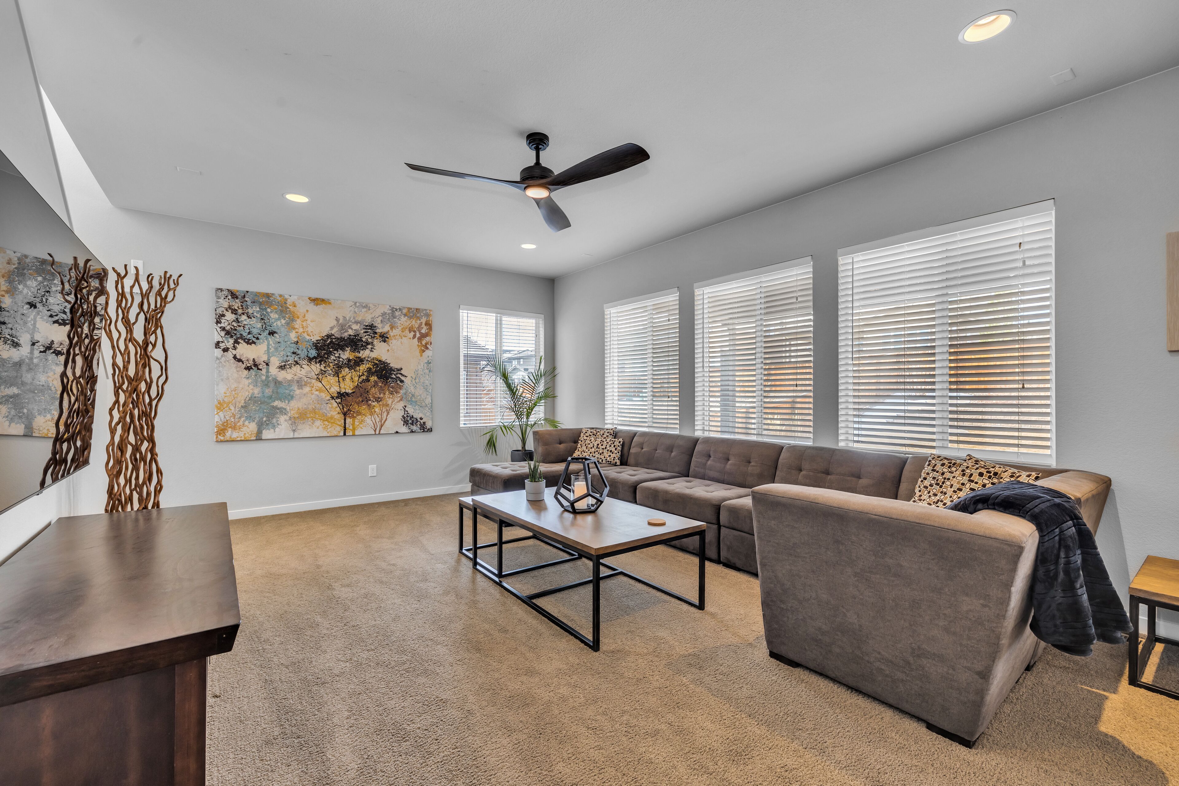 A spacious living room with a gray sofa, a coffee table, and a ceiling fan. There are large windows with blinds and abstract paintings on the walls.
