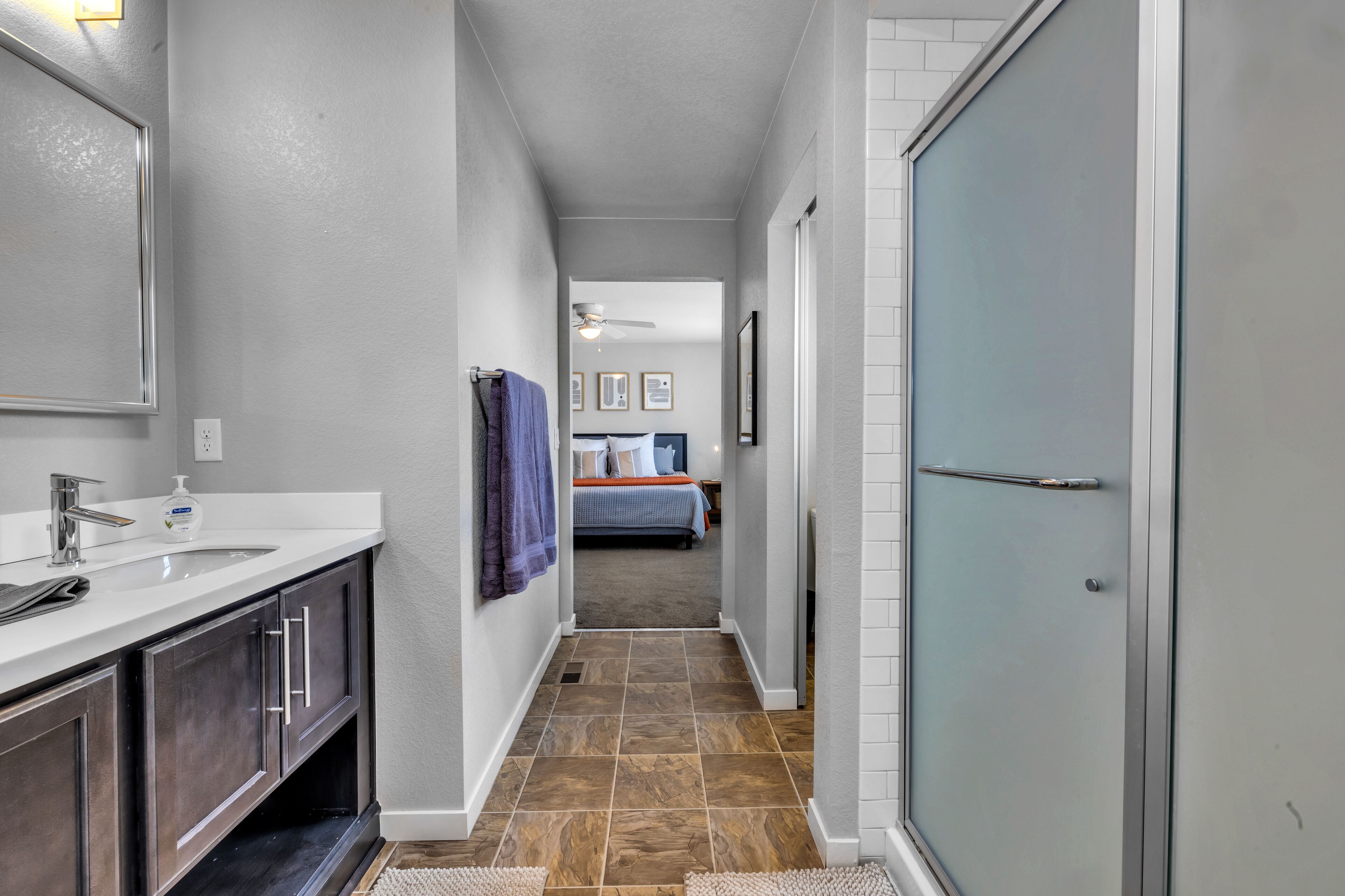 A narrow hallway with a bathroom on the left, a bedroom with a bed and a ceiling fan in the center, and a sliding glass door on the right.