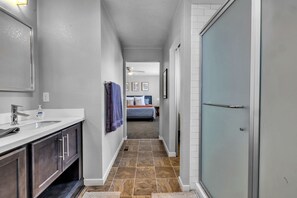 Un couloir étroit, avec une salle de bain sur la gauche, une chambre avec un lit et un ventilateur de plafond au centre, et une porte-fenêtre coulissante sur la droite.