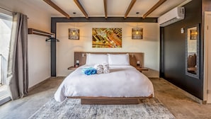 Habitació d'hotel moderna amb un llit gran, bigues de fusta al sostre, aire condicionat fixat a la paret i un quadre abstracte gran sobre el llit.