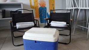 Deux chaises noires avec des coussins blancs, une glacière bleue et blanche, et un tableau coloré accroché au mur.