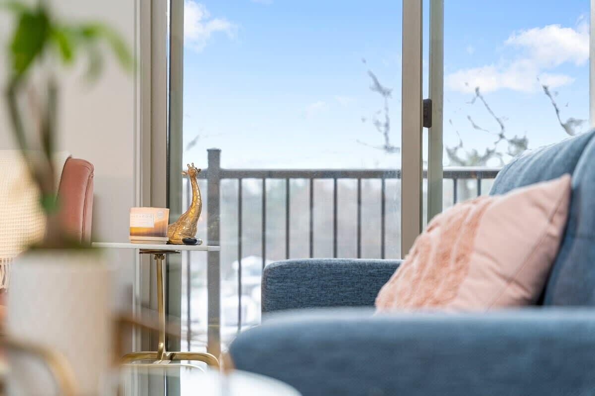 Huoneessa on suuri ikkuna, sininen sohva, jossa on vaaleanpunainen koristetyyny, pieni pöytä, jossa on kynttilä, ja kultainen kirahvihahmo.
